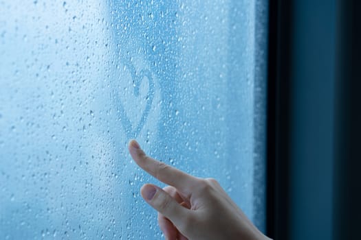 Female hand drawings a heart on a foggy window during the rain. Glass in drops of water. The concept of romance