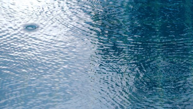 Rain drops on the water pool blue color surface that have ripple wave effect on liquid texture and top view angle. 