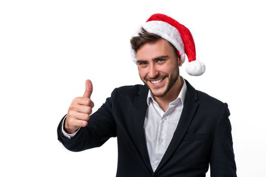 Young handsome caucasian guy in business suit and Santa hats stands on white background in studio smilie and showing thumbs up. Close up portrait business person with Christmas mood Holiday theme