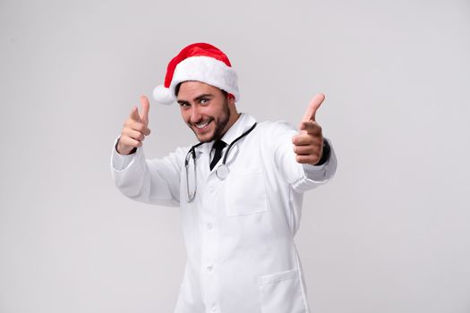 Young handsome doctor in white uniforme and Santa Claus hat standing in studio on white background smile and showing finger in camera Medical student Christmas New Year Holiday Medicine concept