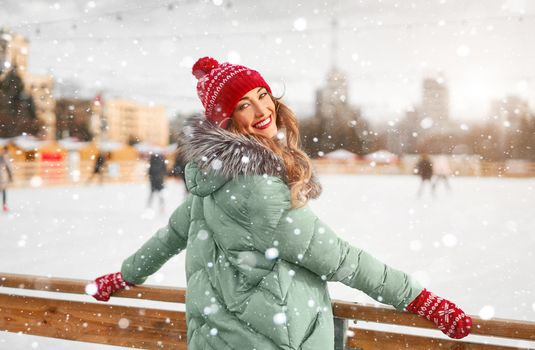 Beautiful lovely middle-aged girl curly hair warm winter jackets red knitted hat glove stands ice rink background Town Square Christmas mood lifestyle Happy holiday woman snowy day Winter leisure