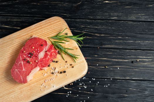 one pieces of juicy raw beef with rosemary on a cutting board on a black wooden table background. Meat for barbecue or grill sprinkled with pepper and salt seasoning