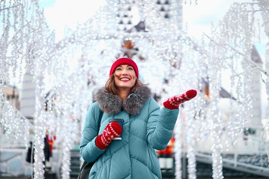 Beautiful lovely middle-aged girl curly hair warm winter jackets red knitted hat glove stands ice rink background Town Square Christmas mood lifestyle Happy holiday woman drink hot beverage coffee