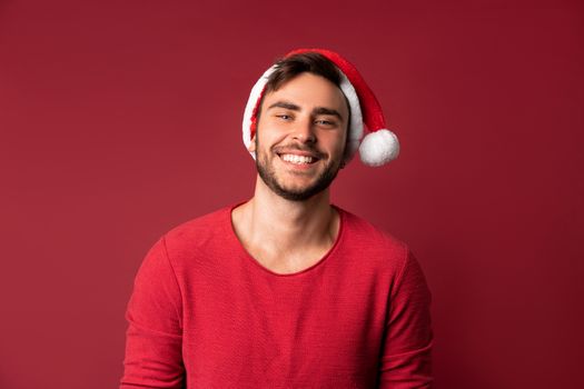 Young handsome caucasian guy in red sweater and Santa hats stands on red background in studio and teeth smiling. Close up portrait european guy with Christmas mood. Holiday theme. New Year.