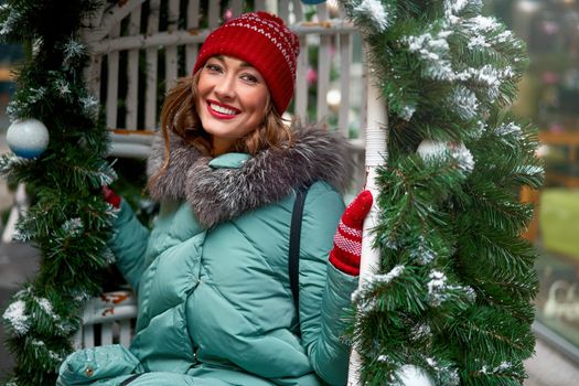 Young beautiful caucasian girl dressed in a warm winter jacket, knitted hat and gloves sits on Christmas decorated swing outdoor Winter holiday concept with happy people smile