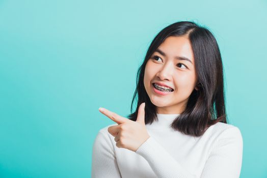 Portrait of Asian teen beautiful young woman smile have dental braces on teeth laughing point finger side away blank copy space, studio shot isolated on blue background, medicine and dentistry concept