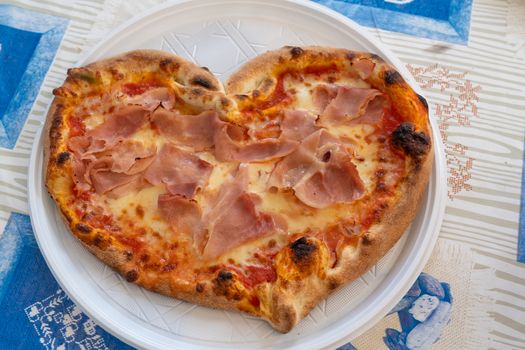 Heart shaped pizza with tomatoes and ham for Valentines Day on white plastic plate on the table,Food concept of romantic love