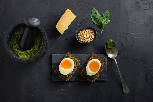 Fresh Green basil pesto in mortar homemade on old rustic wood eggs panini bread with pesto silver spoon on italian breakfast with ingredients green pesto top view on black stone concrete table surface.