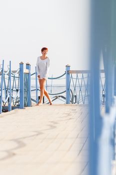 Wide smiling young woman with red short hair cut running on wooden pier. Coastal morning. Natural beauty. Happiness and power of youth.