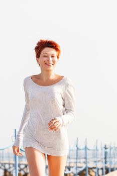 Wide smiling young woman with red short hair cut running on wooden pier. Coastal morning. Natural beauty. Happiness and power of youth.