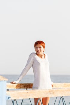 Wide smiling young woman with red short hair cut stays on wooden pier. Coastal morning sunlight. Natural beauty.