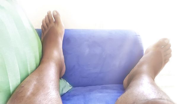 Feet of a bare man resting on a blue sofa with a green pillow. Tiredness and relaxation at home. Rest on the sofa