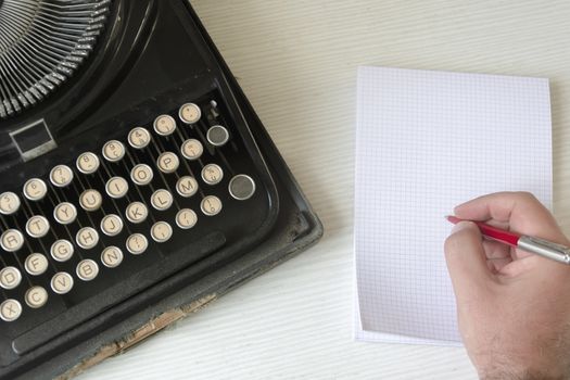 a male hand writing on a white checkered paper with a red pen next to an old wrecked black typewriter. Concept of creativity and storytelling