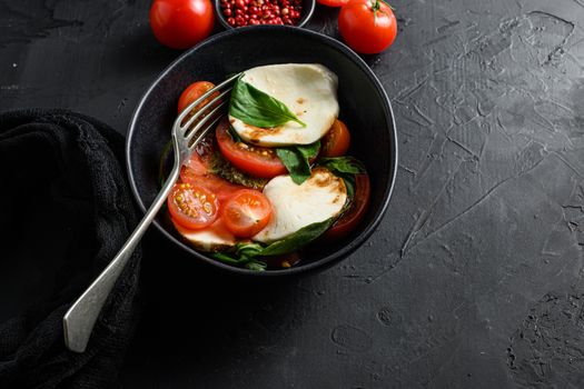Cherry tomatoes, mozzarella buffalo cheese, basil, pesto sauce, and spices on black slate stone chalkboard. Italian traditional caprese salad ingredients. Mediterranean food in black bowl with fork space for text.