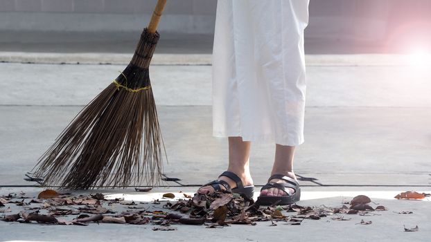 Old woman is sweeping dry leaf on the outdoor cement floor by big long broom.