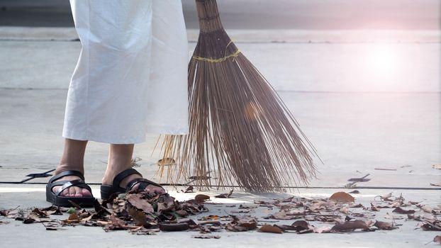 Old woman is sweeping dry leaf on the outdoor cement floor by big long broom.