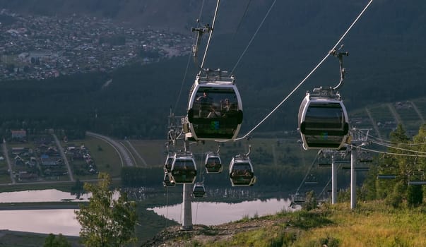 Manzherok resort, Altai Mountains, Russia - August 12, 2020: High angle shot of cableway with tourists inside cable cars on a cityscape and landscape background