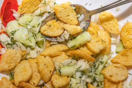 A close up shot of crouton and cucumber salad in a white china plate and table spoon in it.