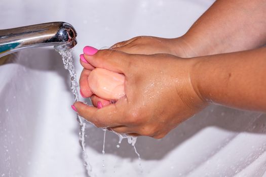 Close up shot of a woman lathering her hands with a soap bar using tap water. Pink finger nails. Hygiene concept.