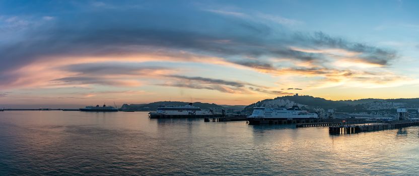 Port of Dover, England - June 24, 2020: Panoramic wide angle shot of Dover Port with P&O, DFDS ferry boats, cruise ship docked at sunset. Beautiful orange, red, blue sky and hills in the background.