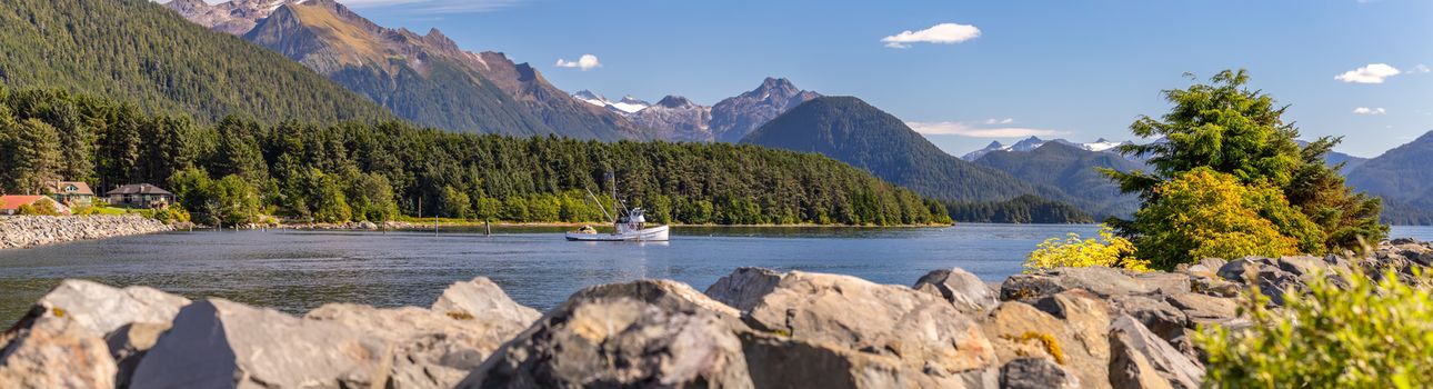 Beautiful panoramic shot of a harbour in Sitka, AK. Commercial fishing boat anchored in it, green forest, mountains with snowy peaks, and gorgeous blue sky. Blurred stone barrier in the foreground.