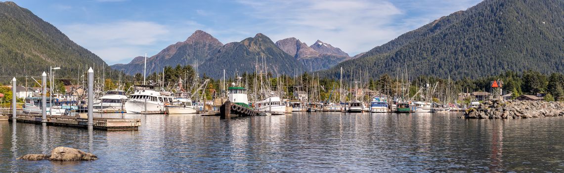 A beautiful panoramic shot of a marina in Sitka, AK with fishing boats, tugboats, yachts docked in it. Green forest, mountains, blue sky in the background. Pier and stone barrier in the foreground.