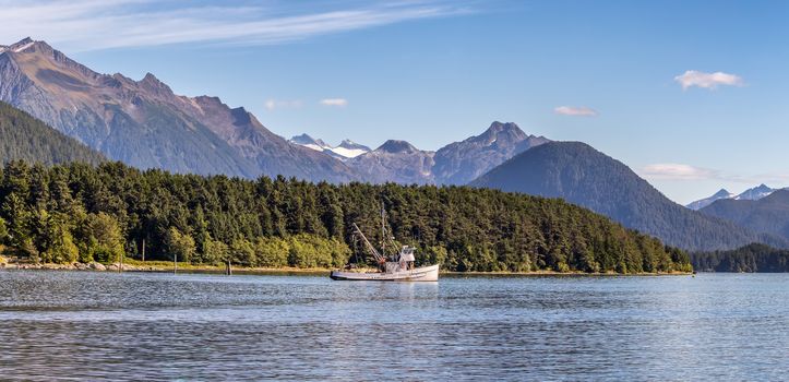 Beautiful panoramic shot of a commercial fishing boat anchored in a harbour in Sitka, AK. Green forest, mountains with snowy peaks, and gorgeous blue sky in the background.