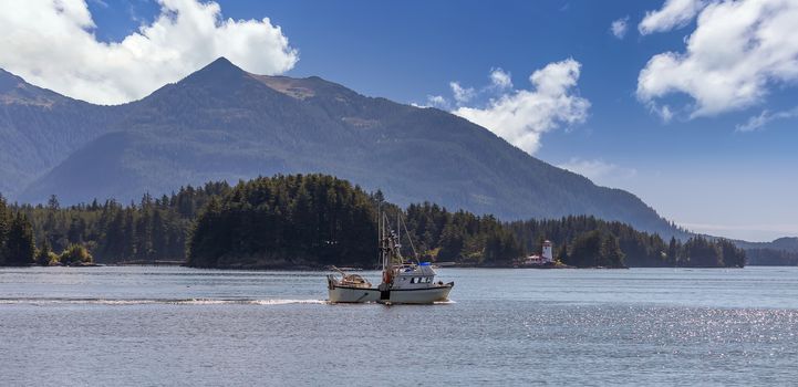 Shot of a commercial fishing boat sailing in a harbour in Sitka, AK. Green forest, mountains, beautiful blue sky with clouds, and a lighthouse in the background.