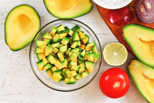 Avocado halves, limes tomato and onion - basic guacamole ingredients on white working board, view from above.