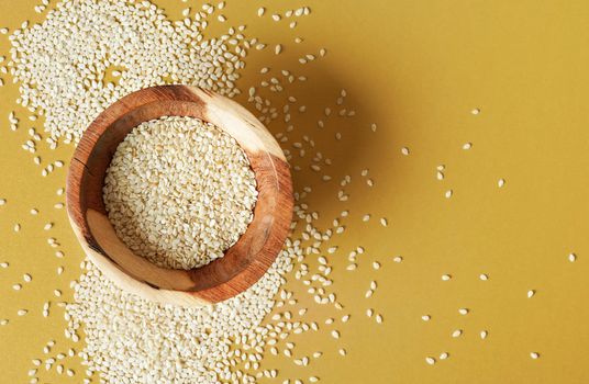 Tiny white sesame seeds in small wooden bowl, on yellow desk, view from above.