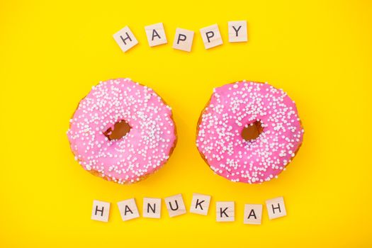 Bright pink delicious donuts on yellow background and inscription from wooden blocks happy hanukkah