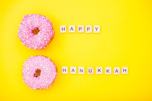Bright pink delicious donuts on yellow background and inscription from wooden blocks happy hanukkah