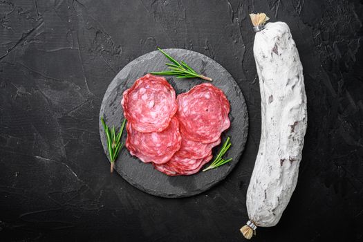 Dry cured, whole and slices cuts of salchichon sausage on black textured surface, topview with space for text.
