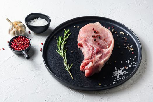 Raw Pork Loin chops in a black round plate on a white stone background with rosemary garlic peppercorns ingredients for grill side view .