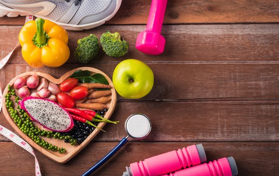 Top view of fresh organic fruit and vegetable in heart plate, shoes, sports equipment and doctor stethoscope, studio shot on wooden gym table, Healthy diet vegetarian food concept, World food day