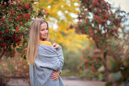 Blonde girl in a gray cardigan and a yellow dress among the autumn trees with red berries. Autumn theme