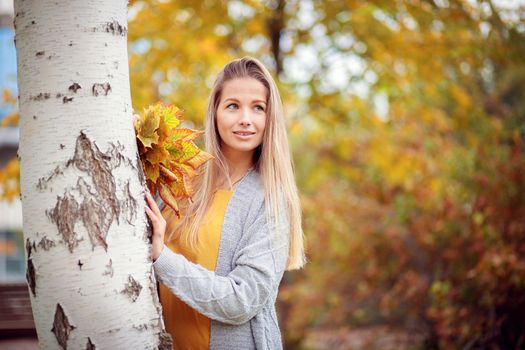 A girl in a gray cardigan and a yellow dress collects autumn leaves in the park