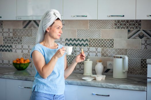 Happy Girl with a towel on her head sutra drinks coffee with cookies for breakfast in his own kitchen