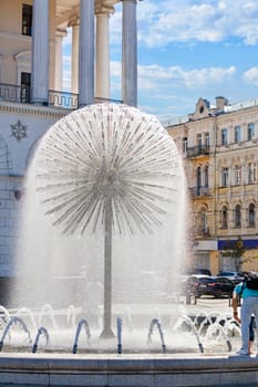 A light refreshing drizzling breeze from the city fountain in the shape of a huge dandelion gives people in the summer heat the freshness of water coolness against the background of the architecture of an old building and a blue slightly cloudy sky.