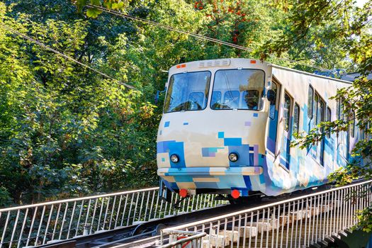 A blue and white cable funicular, illuminated by bright sunlight, rises on rails along the slope, surrounded by green foliage of trees.