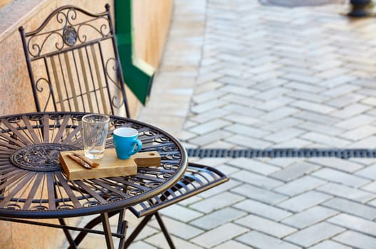 Street still life of a stylish metal table with a turquoise cup, a glass with a metal spoon on a wooden stand, against a background of paving stones in blur, selective focus, text space.