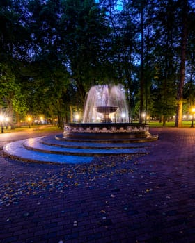 A fountain with blurred streams of water in a night park illuminated by lanterns with a stone pavement, trees and benches. Cityscape.