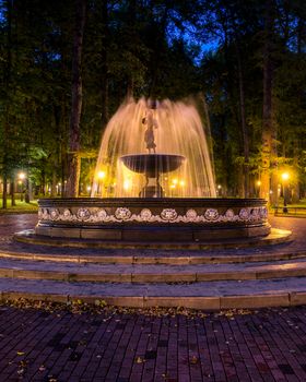 A fountain with blurred streams of water in a night park illuminated by lanterns with a stone pavement, trees and benches. Cityscape.