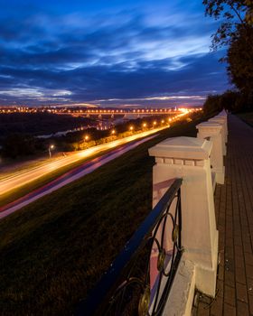 Moving car with blur light through city at night. Bridge over the river and the road. A view from the park from a height with a fence in the foreground. Cityscape.