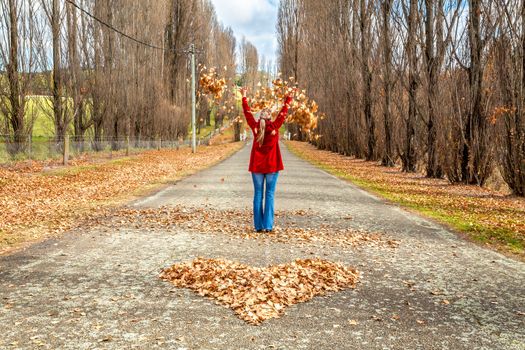 Woman standing in middle of country road, tossing  leaves in air in front of heart of fallen leaves Autumn