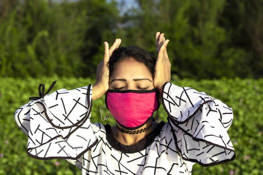 stylish Woman wearing protective pink face mask and posing on natural green background. Stressed Exhausted Young lady Having Strong Tension Headache.