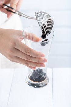 Making bubble tea, scoop and pour cooked brown sugar flavor tapioca pearl bubble balls into cup on white wooden table background, close up, copy space.