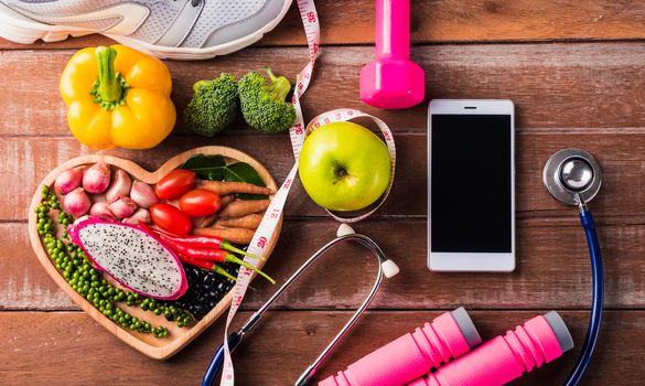 Top view of fresh organic fruit and vegetable in heart plate, shoes, sports equipment and doctor stethoscope, studio shot on wooden gym table, Healthy diet vegetarian food concept, World food day