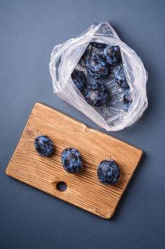 Fresh ripe plum fruits in plastic bag package and on wooden cutting board on minimal blue grey background, top view