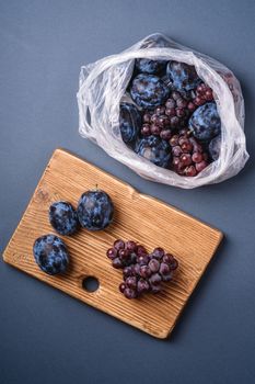 Fresh ripe plum fruits and grape berries in plastic bag package and on wooden cutting board on minimal blue grey background, top view